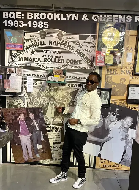 Dr. Glenn Toby at the Universal Hip Hop Museum, standing in front of a wall-sized reproduction of his First Annual Rappers Awards flyer